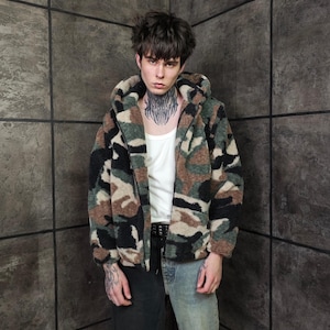 Op de afbeelding: Een camouflage jack met capuchon in groen, bruin en zwart. De jas wordt gedragen over een witte tanktop en gecombineerd met een zwarte broek en een blauwe jeans. De jas heeft een zacht, getextureerd uiterlijk.