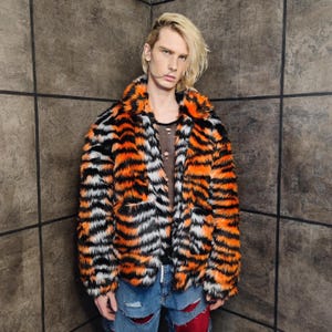 Op de afbeelding: Een faux fur jas met tijgerstrepen in zwart, wit en oranje. De jas heeft een kraag en twee voorzakken. De persoon draagt een ripped blauwe jeans.