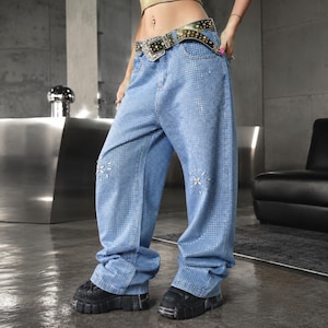 May include: Light blue denim jeans with a loose fit, covered in small, reflective embellishments. The jeans feature a decorative floral design near the knees and are paired with chunky black platform shoes and a studded belt.