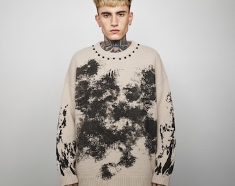 May include: Beige sweater with a black abstract design across the front and sleeves. The sweater has a crew neck with small black studs around the collar. The model is wearing black and white polka dot pants.