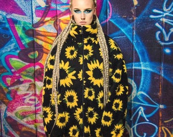May include: A black fleece jacket with a yellow sunflower pattern. The jacket has a high collar and a zipper closure. The background is a colorful graffiti wall.