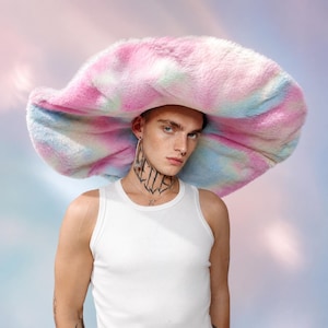 May include: An oversized, wide-brimmed hat made of soft, plush faux fur featuring a tie-dye pattern in shades of pastel pink, blue, and white. The hat has a circular, floppy silhouette with a thick, textured pile that covers the entire crown.