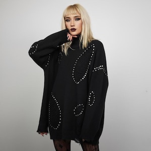 Könnte beinhalten: Schwarzer Oversize-Pullover mit Rollkragen und Distressed-Details. Der Pullover hat silberne Nieten-Verzierungen um die Distressed-Bereiche. Die Ärmel sind lang und der Stil ist lässig.