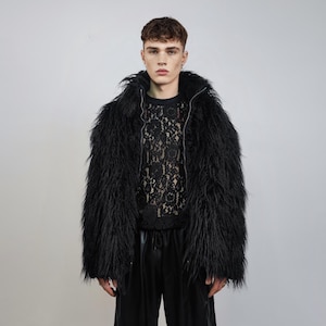 May include: A black faux fur jacket with a zip-up front and a high collar. The jacket has long, shaggy fur and is worn over a black lace top. The model is also wearing black trousers.