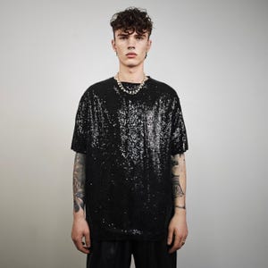 Pode incluir: T-shirt preto com lantejoulas, mangas curtas e gola redonda. A camisa é combinada com calças pretas. Um colar de pérolas é usado. O conjunto é brilhante.