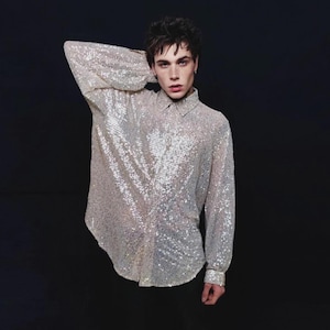 Puede incluir: Una camisa plateada de lentejuelas de manga larga con cuello clásico y puños abotonados. La camisa está hecha de una tela brillante que refleja la luz, creando un efecto deslumbrante. La camisa se usa sobre un fondo oscuro.
