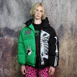 May include: A two-tone puffer jacket, half green and half black, with a hood. The green side features the letters "BM" and a yin-yang flame design. The black side displays a white skeleton print. The jacket is worn with a black shirt and pink patterned pants.