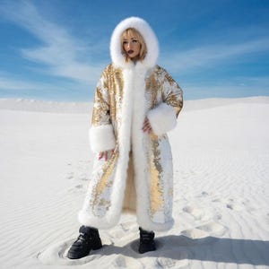 May include: A long, hooded coat with gold sequins and white faux fur trim. The coat features a white fur-lined hood, cuffs, and front trim. The person is wearing black boots. The background is a white desert under a blue sky.