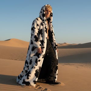 May include: A long, hooded coat featuring a black and white cow print design. The coat is set against a desert backdrop, with sand dunes visible. The coat's material appears to be a soft, faux fur.