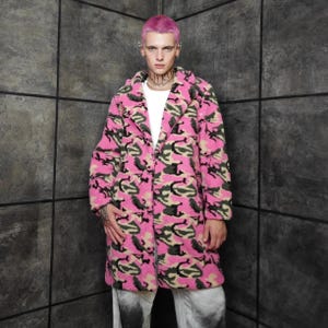 Könnte beinhalten: Ein rosa Camouflage-Mantel mit klassischem Reverskragen. Der Mantel hat ein Muster aus grünen und schwarzen Formen auf einem hellrosa Hintergrund. Der Mantel wird über einem weißen T-Shirt getragen.