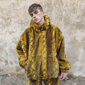 Może przedstawiać: Kurtka z kapturem i pasujące spodnie z imitacji futra z żółto-brązowym wzorem wężowej skóry. Kurtka ma zapięcie na zamek błyskawiczny i kieszenie. Strój wykonany jest z miękkiego, pluszowego materiału.