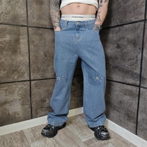 May include: Light blue denim jeans with a wide leg fit, covered in small, sparkling embellishments. The jeans feature decorative stitching near the knees. The person is wearing black and gray platform shoes.