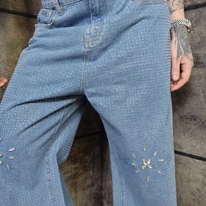 May include: Light blue denim jeans with a pattern of small, silver-colored studs covering the entire surface. The jeans have a classic five-pocket design and feature decorative rhinestone accents near the knees. The jeans are a wide-leg style.