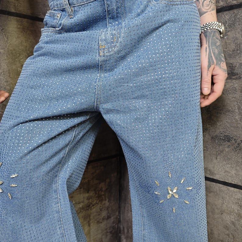 May include: Light blue denim jeans with a pattern of small, silver-colored studs covering the entire surface. The jeans have a classic five-pocket design and feature decorative rhinestone accents near the knees. The jeans are a wide-leg style.