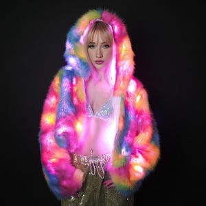 May include: A vibrant, rainbow-colored faux fur hooded jacket with integrated LED lights. The jacket features a mix of pink, yellow, blue, and orange hues, creating a striking visual effect. The design includes a hood and a cropped silhouette, perfect for festivals or parties.