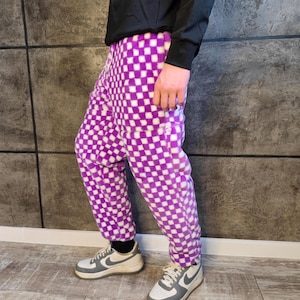 Peut inclure: Pantalon en polaire à carreaux violets et blancs. Le pantalon a une coupe ample et une texture douce. La personne porte des baskets grises et blanches avec des chaussettes noires et un haut noir à manches longues.