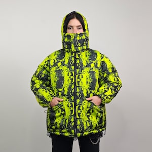 Può includere: Una giacca imbottita di colore giallo brillante con una stampa astratta in bianco e nero. La giacca ha un cappuccio e una chiusura a zip.