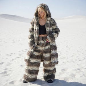 May include: A full-length faux fur coat and matching pants set in a gray and white spotted pattern. The coat has a hood and the pants are wide-legged. The outfit is worn in a desert setting.