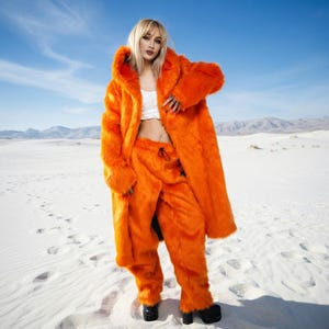 Può includere: Un accattivante completo composto da cappotto e pantaloni in finta pelliccia arancione. Il cappotto lungo ha un cappuccio e i pantaloni hanno una vita con coulisse. L'outfit è indossato con un top corto bianco e scarpe nere. Lo sfondo è un deserto bianco sotto un cielo blu.
