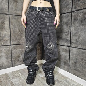 May include: Black pants with a small white dot pattern and decorative floral designs on the knees. The pants have a black belt with a silver buckle and are paired with black platform boots. The pants are a loose fit.