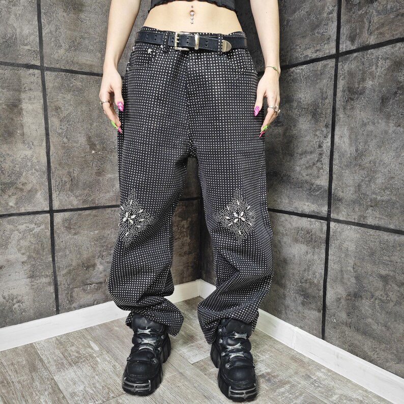 May include: Black pants with a small white dot pattern and decorative floral designs on the knees. The pants have a black belt with a silver buckle and are paired with black platform boots. The pants are a loose fit.