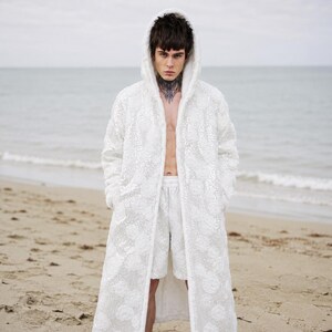 May include: A white hooded coat and matching shorts with a floral pattern. The coat is open, revealing the chest. The outfit is worn on a sandy beach with the ocean in the background.