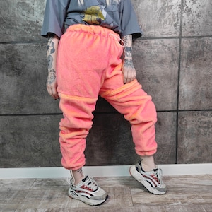 Könnte beinhalten: Helle pinkfarbene Fleece-Jogginghose mit elastischem Bund und Kordelzug. Die Hose hat einen kontrastierenden orangefarbenen Besatz an den Bündchen und am Bund. Das Model trägt weiße, graue und rote Turnschuhe.