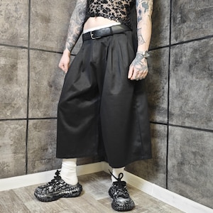 May include: Black culottes with pleats, paired with a black belt and sneakers. The wide-leg pants are made of a solid, dark fabric. The sneakers have a black and white design.