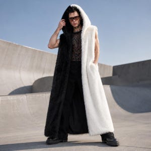 May include: A long, sleeveless faux fur coat in a striking black and white split design. The coat features a large hood and pockets. The model is wearing black pants and boots. The coat's length is floor-length.