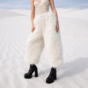 Può includere: Un top a corsetto color crema e pantaloni bianchi e soffici sono abbinati a stivali con plateau neri. I pantaloni hanno una consistenza pelosa. Gli stivali hanno un tacco grosso. Lo sfondo è un paesaggio desertico bianco.