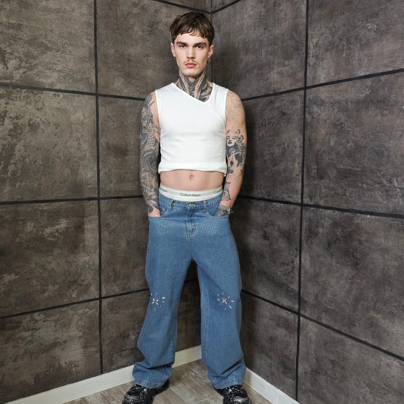 May include: A white sleeveless top and light blue denim jeans with star-shaped embellishments. The jeans have a wide leg design. The model is wearing black shoes and Calvin Klein underwear.