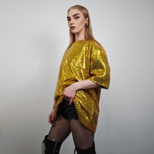 Könnte beinhalten: Ein goldfarbenes Pailletten-Oberteil mit kurzen Ärmeln. Das Oberteil ist locker geschnitten und bedeckt den Oberkörper. Das Model trägt schwarze Shorts, Netzstrümpfe und schwarze Stiefel. Der Hintergrund ist eine schlichte weiße Wand.