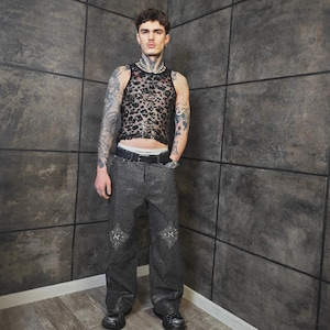 May include: A black, see-through, leopard-print tank top and black studded pants with decorative patches. The pants have a black belt with a silver buckle. The model is wearing black shoes.