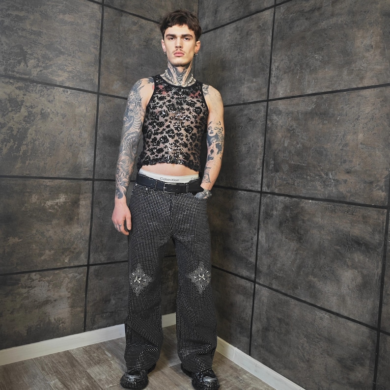 May include: A black, see-through, leopard-print tank top and black studded pants with decorative patches. The pants have a black belt with a silver buckle. The model is wearing black shoes.