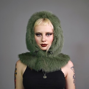 May include: A green faux fur hooded scarf with a drawstring closure. The scarf is worn by a person with blonde hair and dark makeup.