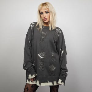 May include: A dark grey distressed jumper with large ripped sections. The jumper has a ribbed hem and cuffs, with a white trim at the bottom. The sleeves are long, and the overall style is oversized.