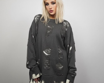May include: A dark gray distressed sweater with large ripped sections. The sweater has a ribbed hem and cuffs, with a white trim at the bottom. The sleeves are long, and the overall style is oversized.