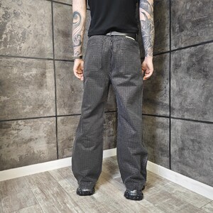May include: Black and silver patterned pants with a grid-like design. The pants have a relaxed fit and are worn with black shoes. The image is taken from a low angle, showing the full length of the pants.