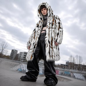 Op de afbeelding: Een lange imitatiebontjas met capuchon en een meerkleurig patroon van witte, zwarte en bruine verticale strepen. De jas wordt gedragen over een zwarte mesh crop top en een wijde zwarte broek met riem, gecombineerd met zwarte plateauzolen.
