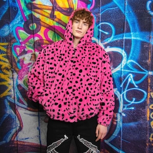 Może przedstawiać: Żywa różowa kurtka z kapturem i czarnymi plamami, przypominająca wzór lamparta. Kurtka ma zapięcie na zamek i jest noszona z czarnymi spodniami. Tło przedstawia kolorowe graffiti.