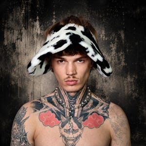May include: A wide-brimmed bucket hat featuring a black and white cow print pattern made of a soft, plush faux fur material. The hat has a rounded crown and a downward-sloping brim that frames the face, providing a distinct textured appearance.