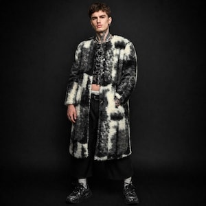 Könnte beinhalten: Ein langer, offener Mantel mit einem schwarz-weißen Muster. Der Mantel hat eine zottelige Textur und ein knielanges Design. Die Person auf dem Bild trägt eine schwarze Hose und Schuhe. Der Hintergrund ist einfarbig schwarz.