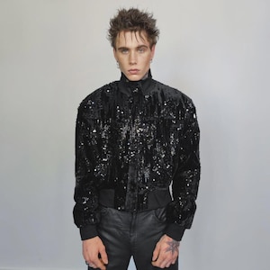 Könnte beinhalten: Schwarze Pailletten-Bomberjacke mit hohem Kragen und gerippten Bündchen. Die Jacke hat eine samtige Textur und wird mit einer schwarzen Hose kombiniert. Der Gesamtstil ist edgy und modisch.
