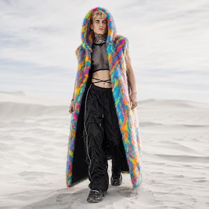 May include: A long, sleeveless coat with a hood, featuring a rainbow of colours. The coat is made of faux fur. The person is wearing black trousers and boots. The background is a sandy landscape.