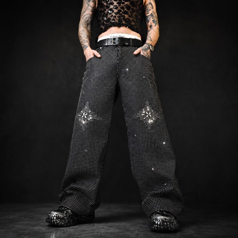 May include: Black, wide-leg pants covered in small, sparkling embellishments. The pants feature decorative, star-shaped rhinestone designs on the front. The pants have a black belt with a silver buckle. The person is wearing black shoes.