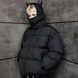 Könnte beinhalten: Schwarze Pufferjacke mit Kapuze, kleinen Hörnern und Gesichtsbedeckung. Die Jacke hat ein auf die Brust gesticktes Totenkopf-Design. Die Jacke ist einfarbig dunkel.