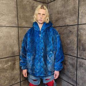 Peut inclure: Veste à capuche en fausse fourrure bleue avec un motif peau de serpent. La veste a une fermeture à demi-zip et une coupe ample. La veste est d'un bleu vif avec des accents bleu foncé et noirs. Le modèle porte un jean déchiré.