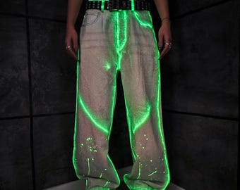 May include: Light-wash denim jeans with a neon green glow effect. The jeans have a relaxed fit, a black belt, and white paint splatters. The neon green outlines the seams and pockets.