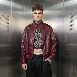 May include: A burgundy faux leather jacket with a snakeskin pattern. The jacket has a high collar and a zipper closure. The model is wearing a sequined top and black pants. The jacket is a bold fashion statement.