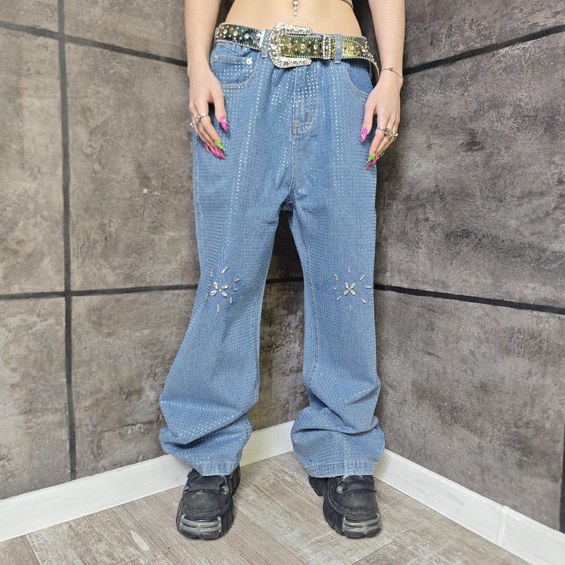May include: Light blue denim jeans with a flared leg, embellished with small, sparkling accents and starburst designs on the knees. The jeans have a high waist and are paired with a decorative belt and platform shoes.